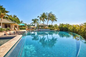 A clear, infinity pool with lounge chairs and a gazebo, surrounded by palm trees and a clear sky.
