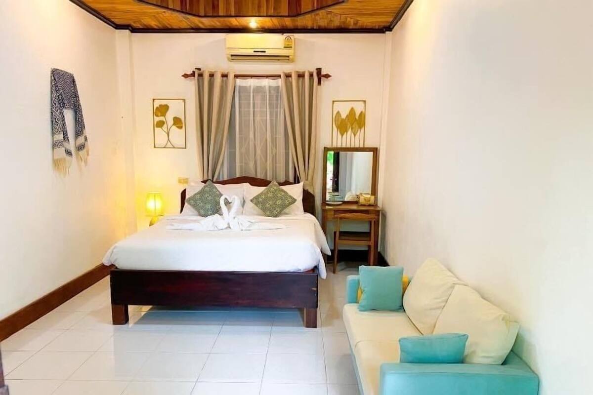 A hotel room with a bed, a wooden headboard, a bedside table with a lamp, a mirror, and a sofa.