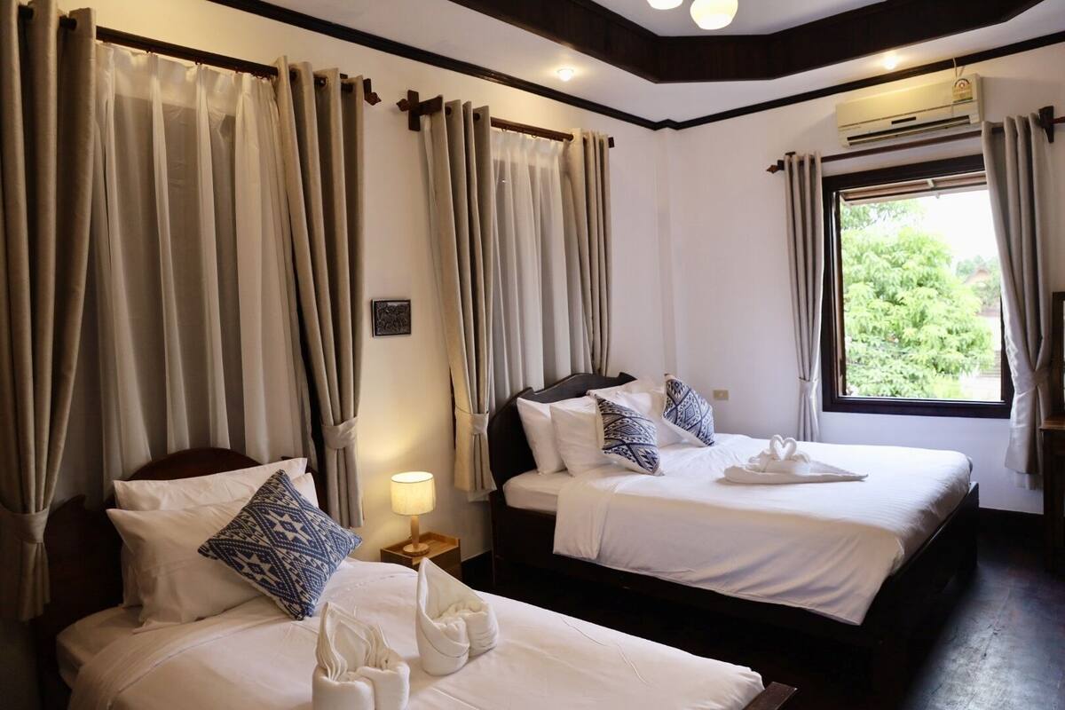 A hotel room with two beds, beige curtains, a bedside lamp, and a window with a view of greenery.