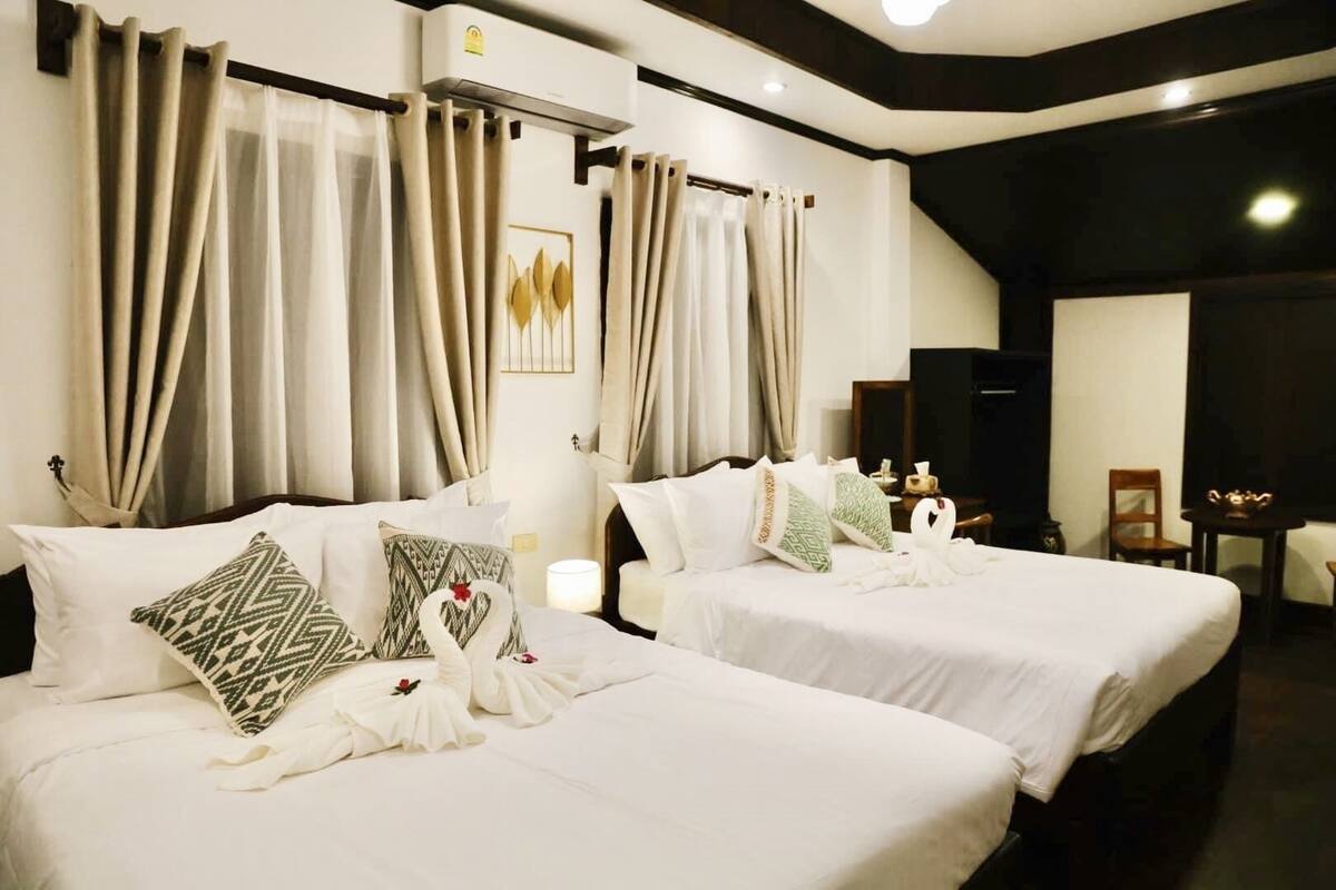 A hotel room with two beds, beige curtains, a wall-mounted air conditioner, and a small table with a lamp.
