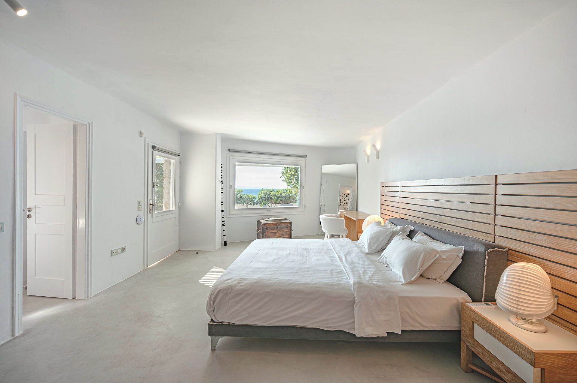 A bedroom with a large bed, a wooden headboard, a bedside table with a lamp, and a view of the outdoors through a window.