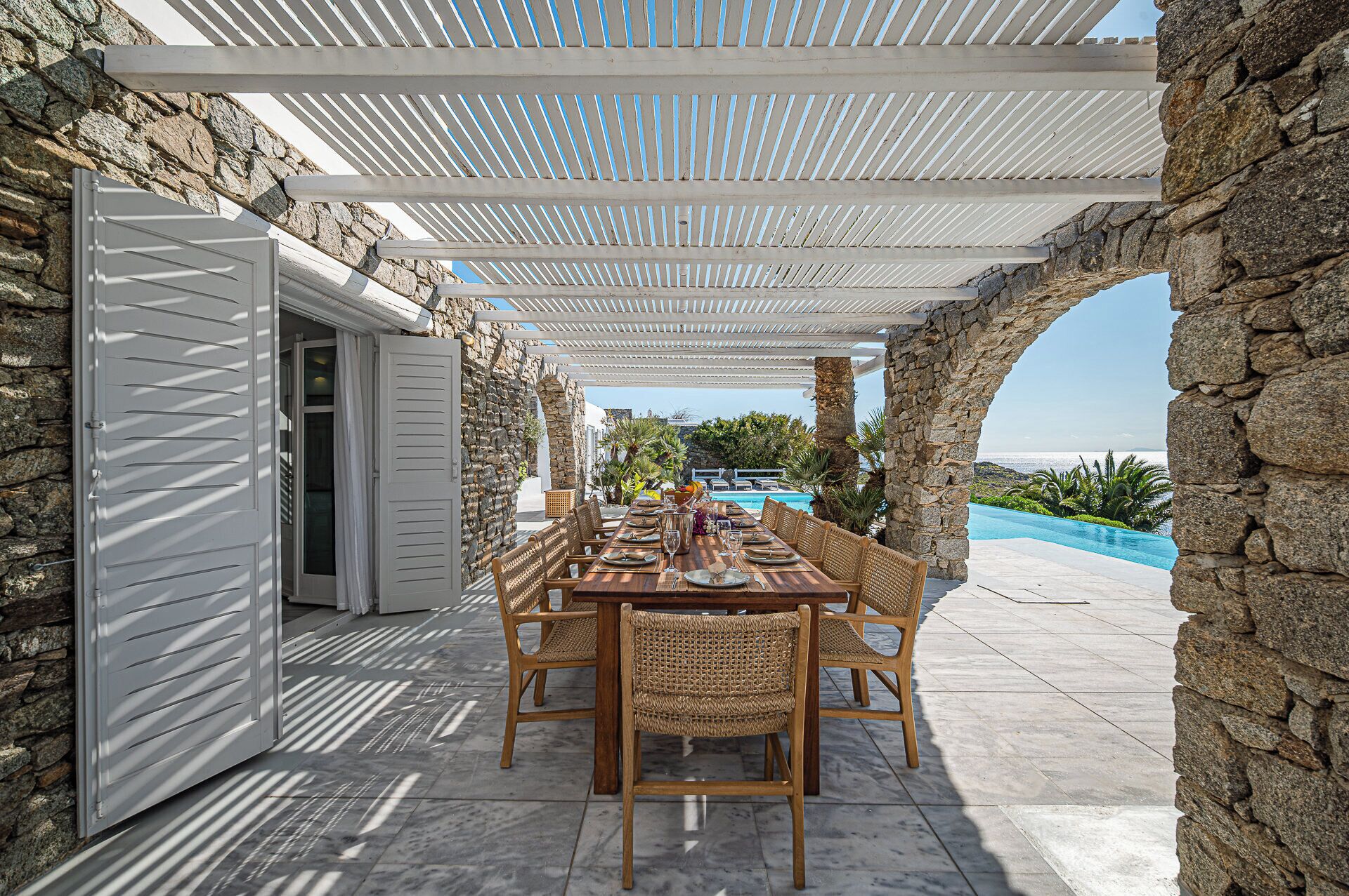 Área de refeições ao ar livre com parede de pedra, pérgola e vista para piscina e paisagem.