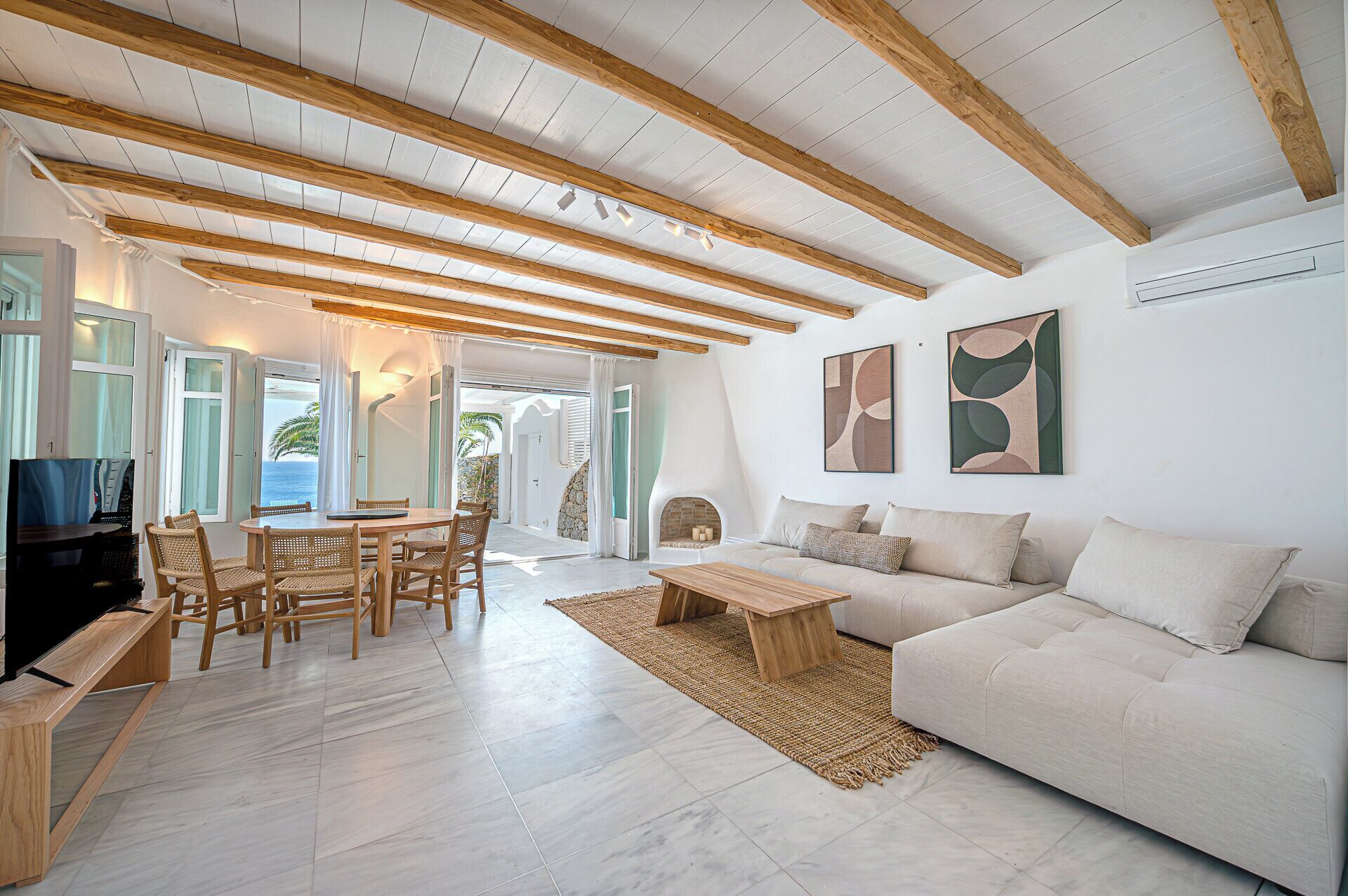 A spacious living area with a wooden ceiling, a white sofa, a dining table with chairs, and a view of the ocean through open doors.
