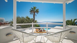 A covered outdoor seating area with two white chairs, a wooden table with food and drinks, and a view of the sea and palm trees. - Sanarte villa 2, 5br w/private pool and sea view! (Mykonos)