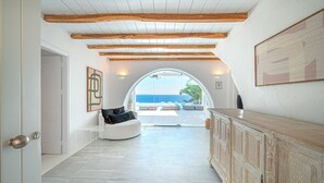 A spacious hallway with a high ceiling, wooden beams, a white sofa with cushions, a large arched doorway leading to a balcony with a view of the sea, and a wooden cabinet. - Sanarte villa 2, 5br w/private pool and sea view! (Mykonos)