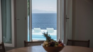 A dining table with a fruit bowl in the center, overlooking a pool with two white lounge chairs and a view of the sea and mountains. - Sanarte villa 2, 5br w/private pool and sea view! (Mykonos)