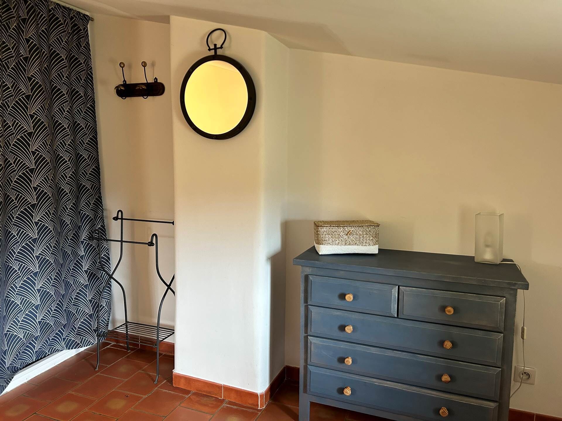 Ein Zimmer mit weißer Wand, einer blauen Kommode mit Schubladen, einem runden Spiegel und einem gemusterten Vorhang.
