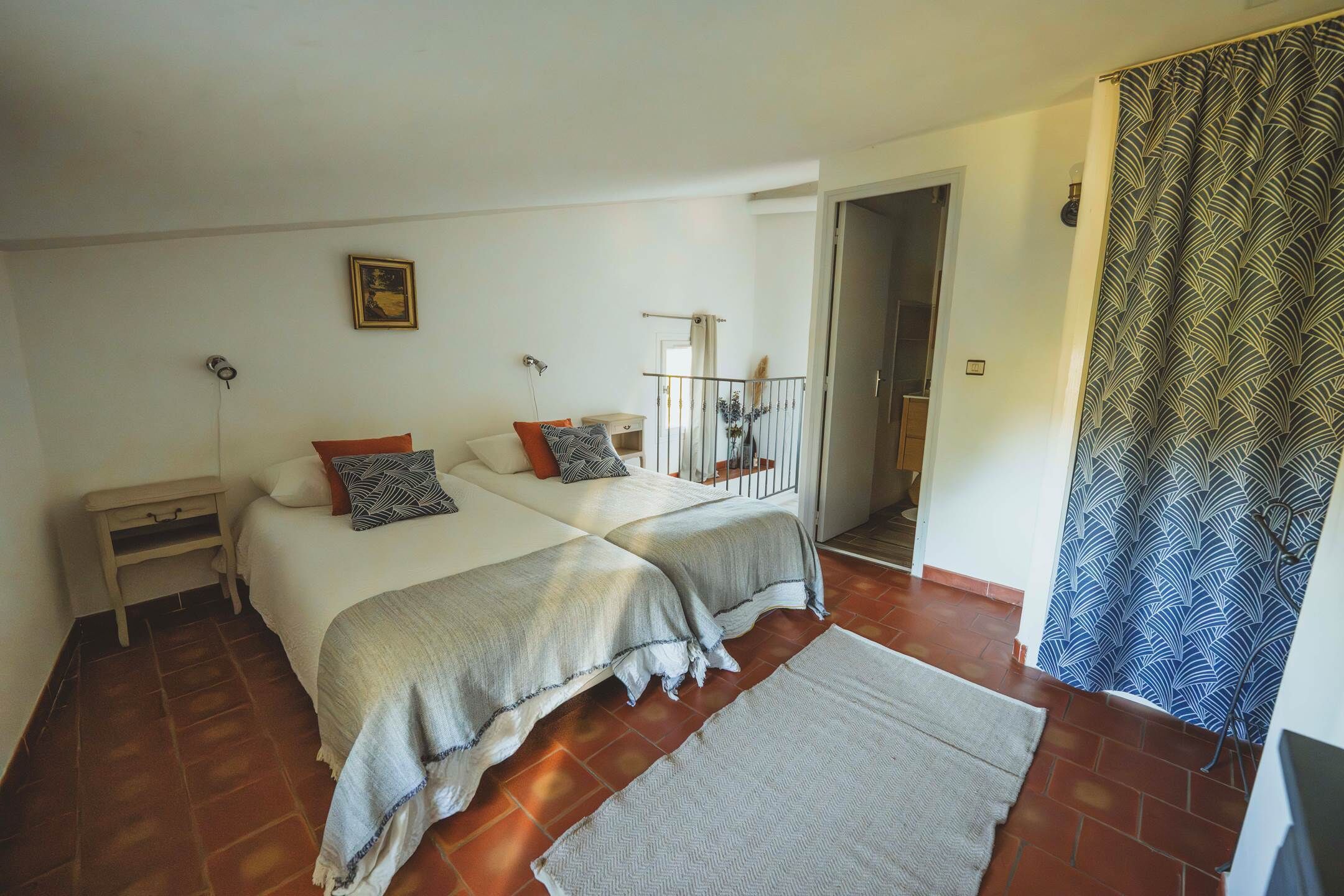 Une chambre avec deux lits, une petite table de chevet, une commode, un rideau à motifs et une vue sur un balcon.