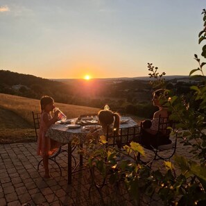 An outdoor dining table with chairs, surrounded by greenery, with a view of a sunset over a hilly landscape. - Santoline Holiday Cottage (Niozelles)