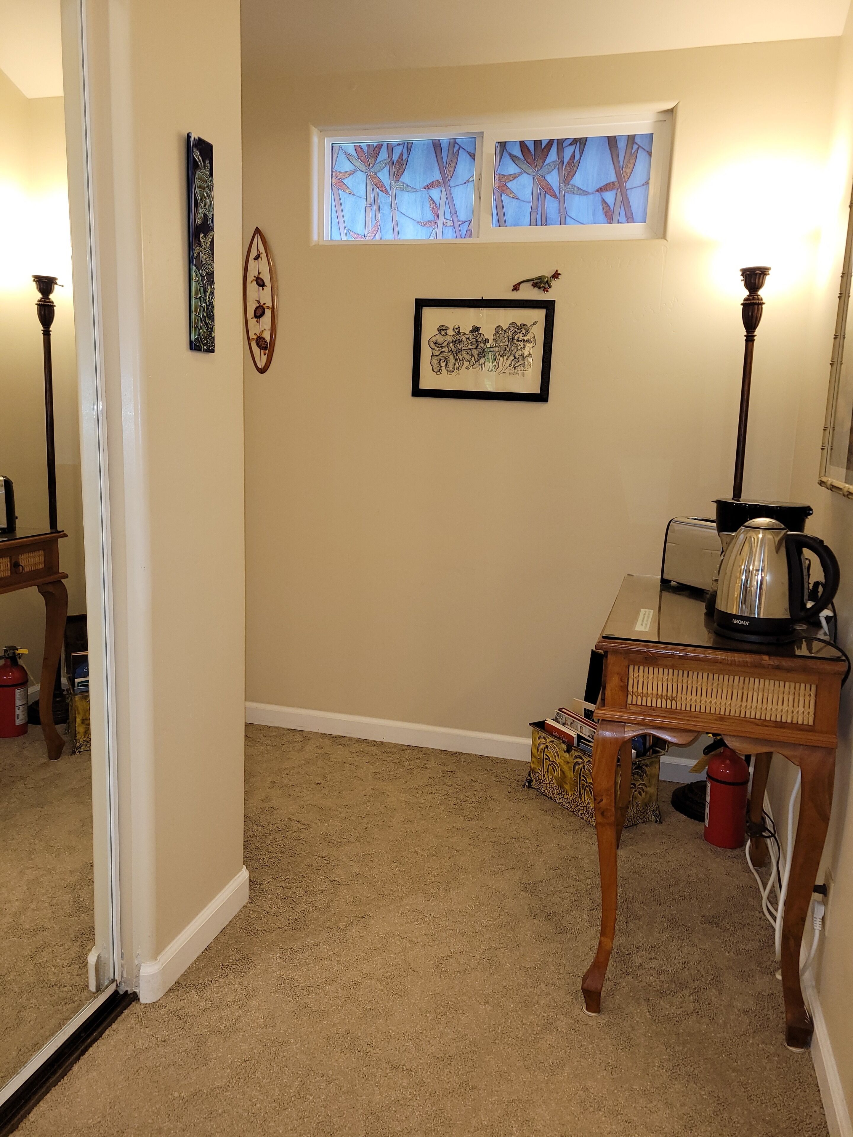 A room with a beige carpet, a wooden table with a kettle, a framed picture, and a window with bamboo artwork.