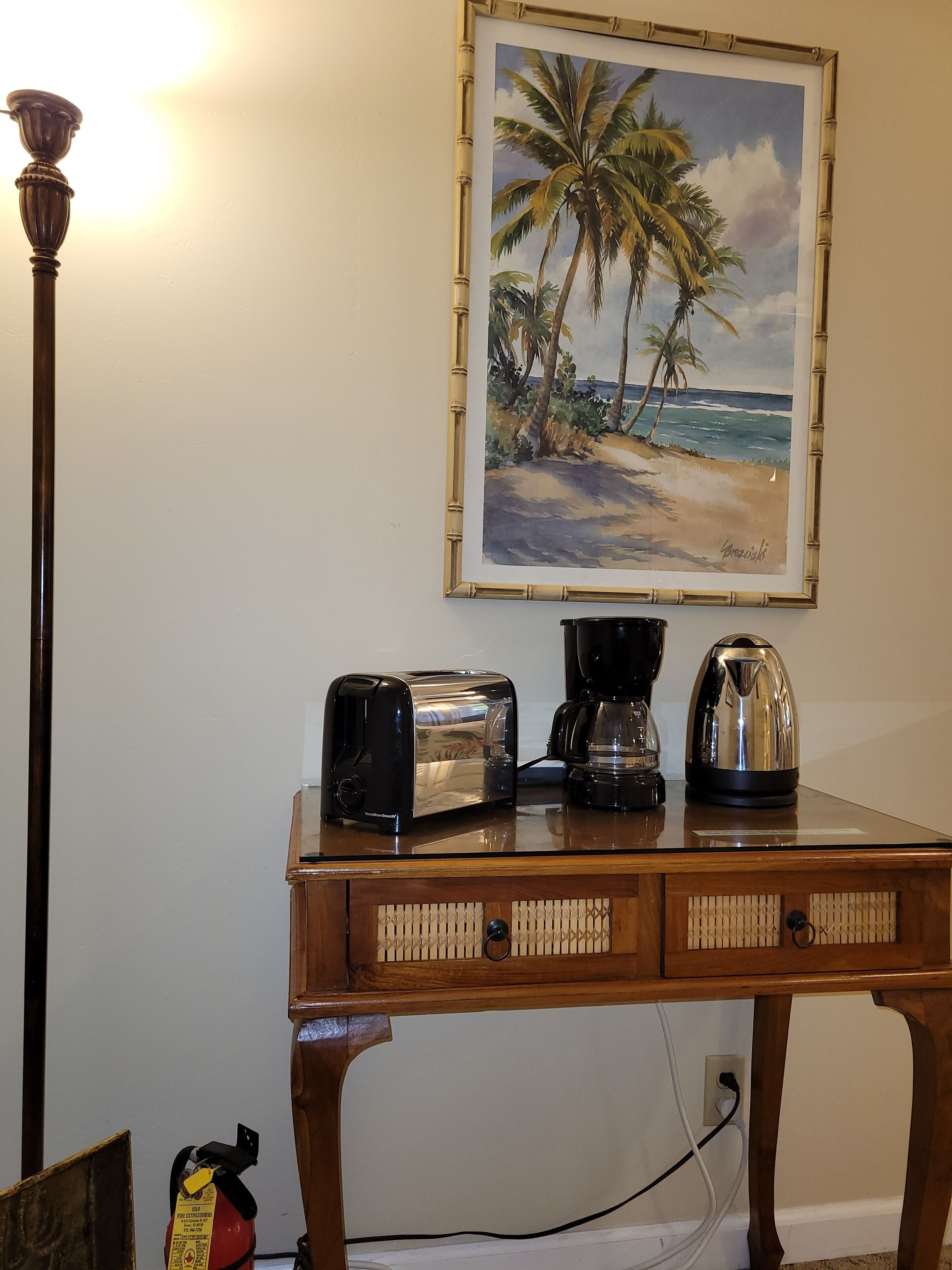 A wooden table with a toaster, coffee maker, and kettle. A fire extinguisher is on the floor. A painting of a beach scene hangs on the wall.