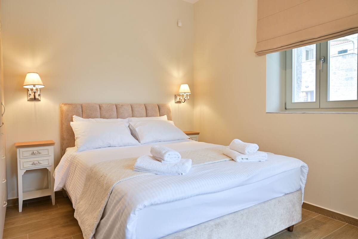 A neatly made bed with white linens and towels, two bedside tables with lamps, and a window with a view of buildings.