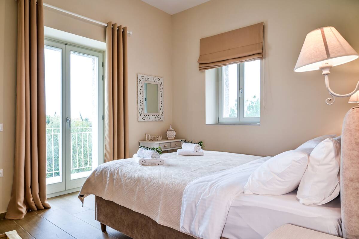 A bedroom with a bed, a window with curtains, a bedside lamp, a mirror, and a dresser with towels.