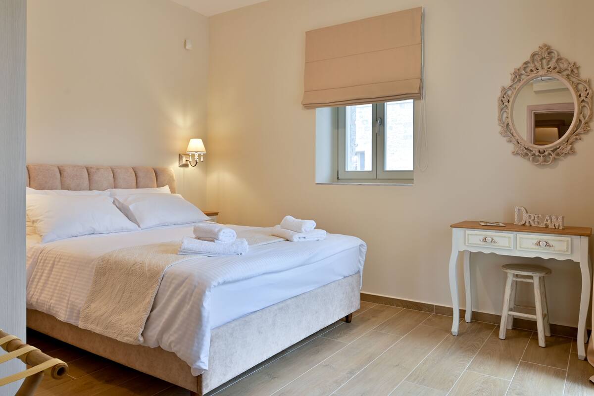 A bedroom with a bed, a desk, a chair, a window with a blind, and a mirror on the wall.
