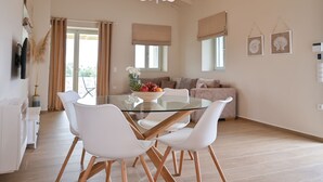 Uma sala de jantar moderna com mesa de vidro e cadeiras brancas, um sofá ao fundo e um lustre.