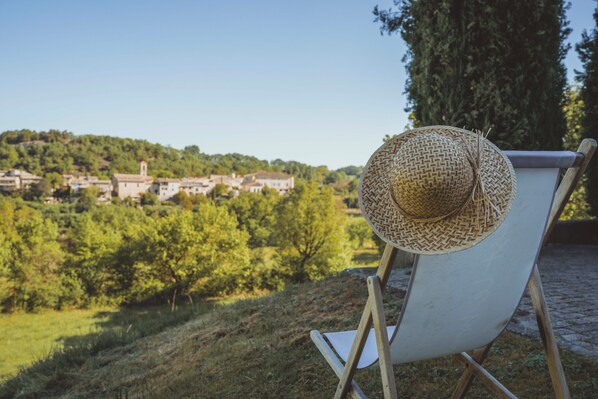 Une chaise surmontée d’un chapeau tressé, une vue dégagée sur un village et un grand arbre en arrière-plan.