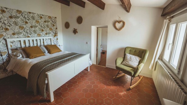 A bedroom with a bed, a rocking chair, a wall with a floral pattern, and a door leading to another room. - Gîte Lavande (Niozelles)
