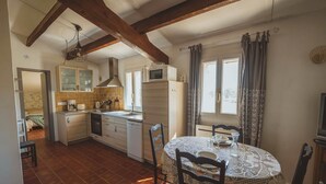 A compact kitchen with a dining area, wooden beams, a tiled floor, and a window with curtains. - Gîte Lavande (Niozelles)
