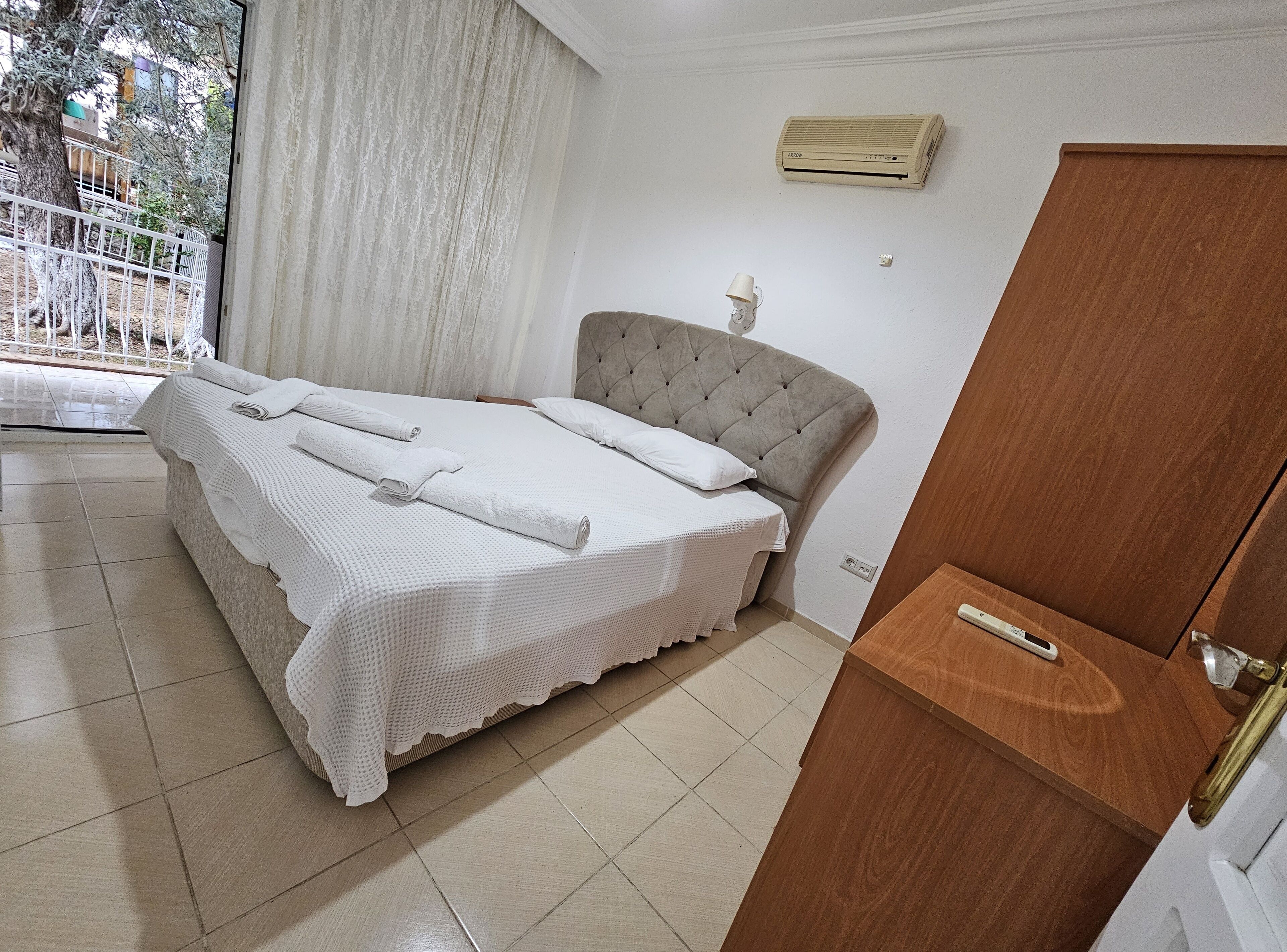 A bedroom with a bed, a wooden nightstand, a window with curtains, and an air conditioning unit on the wall.