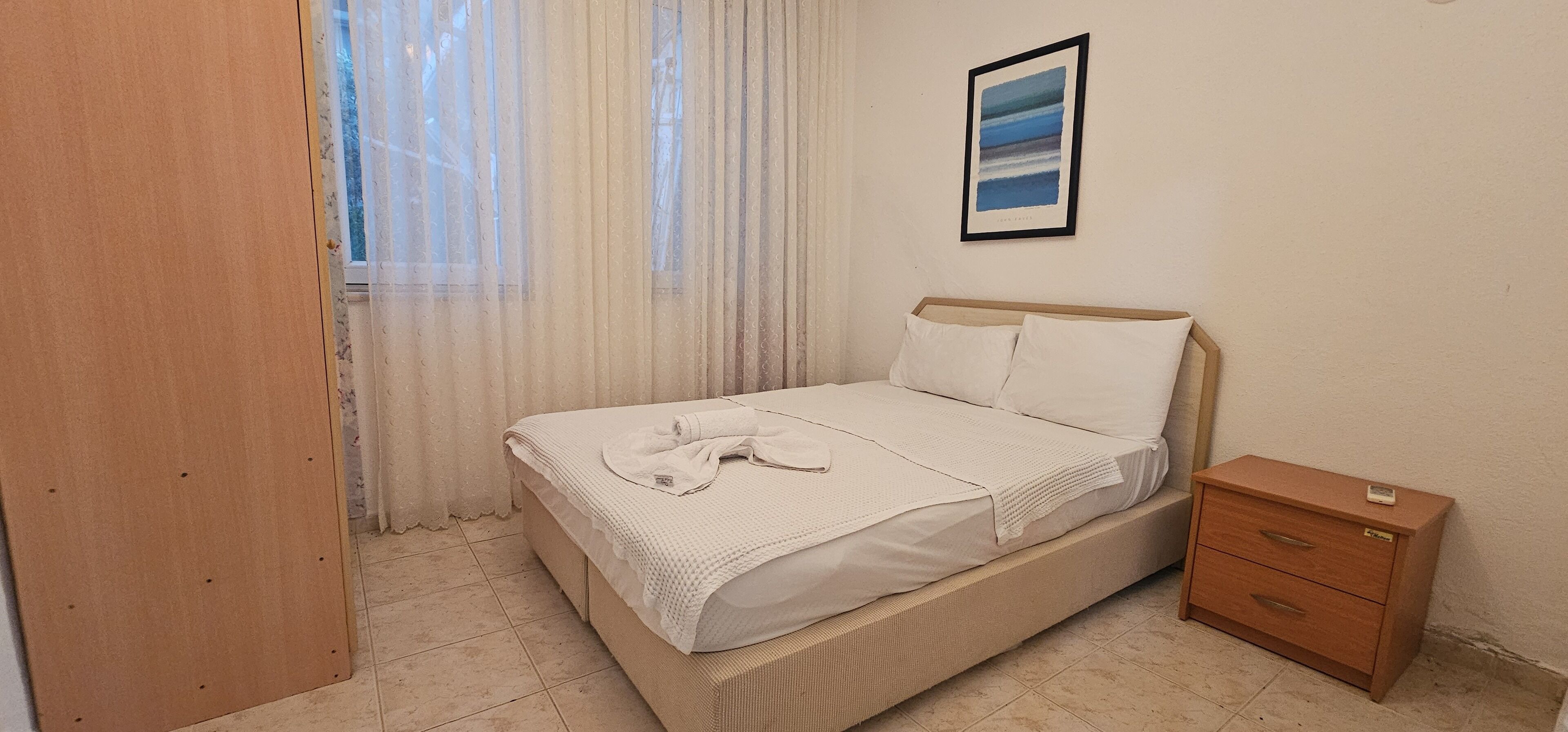 A neatly made bed with a white comforter and pillows, a wooden nightstand with a drawer, a framed picture on the wall, and a window with sheer curtains.
