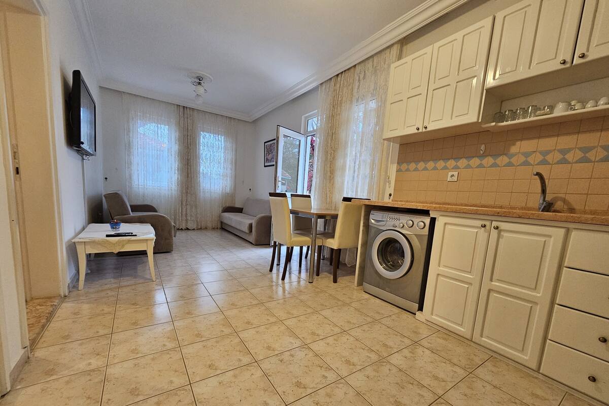 A compact living space with a dining area, a sofa, a washing machine, and a kitchen with white cabinets and a tiled backsplash.