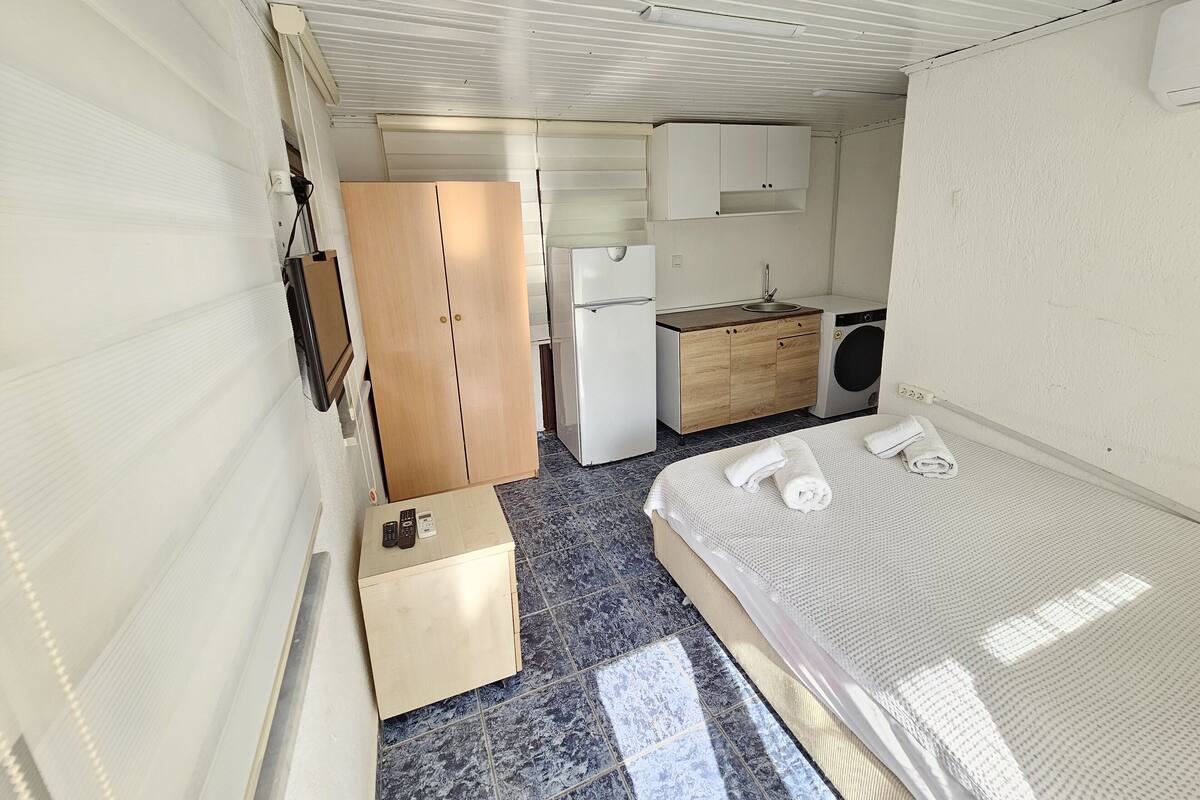 A compact room with a bed, a small kitchenette, a wooden wardrobe, and a washing machine.