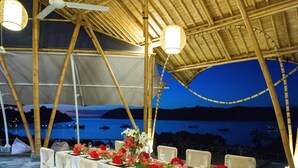 A dining area with bamboo chairs and tables, set up for a meal with a view of the sea and boats in the background. - The Seraya Resort Komodo (Labuan Bajo)