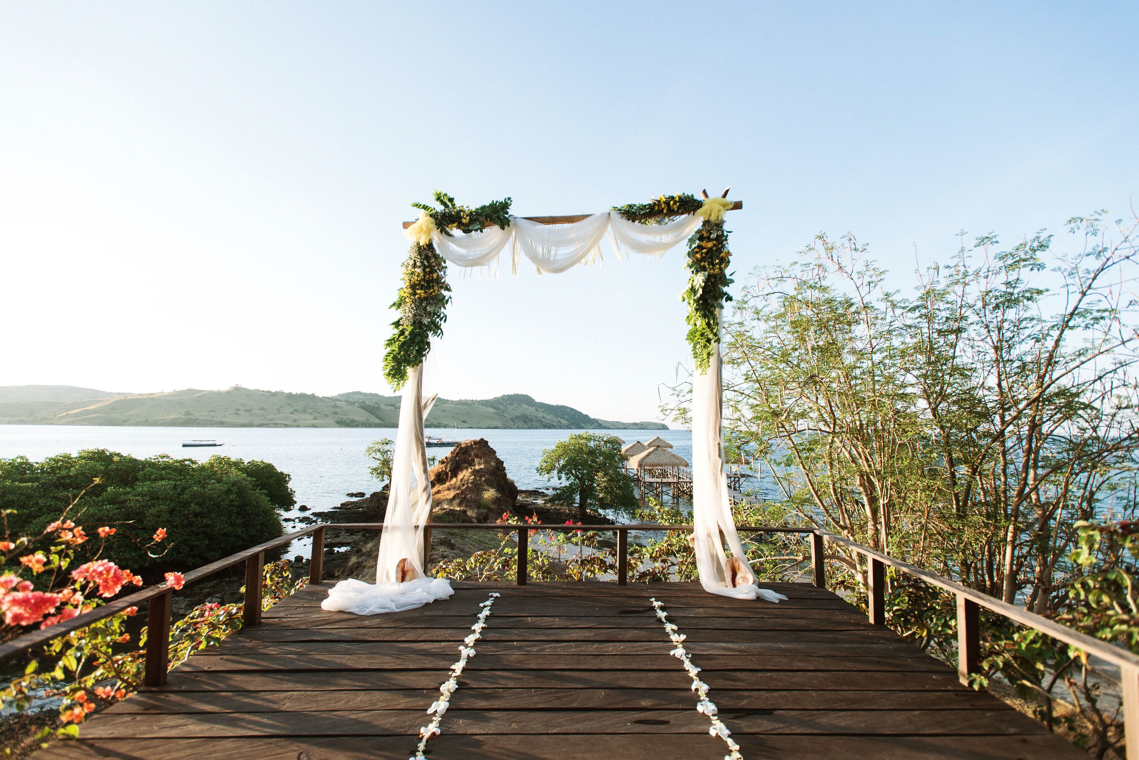 A wooden deck leading to a white arch adorned with greenery, overlooking a body of water and hills.