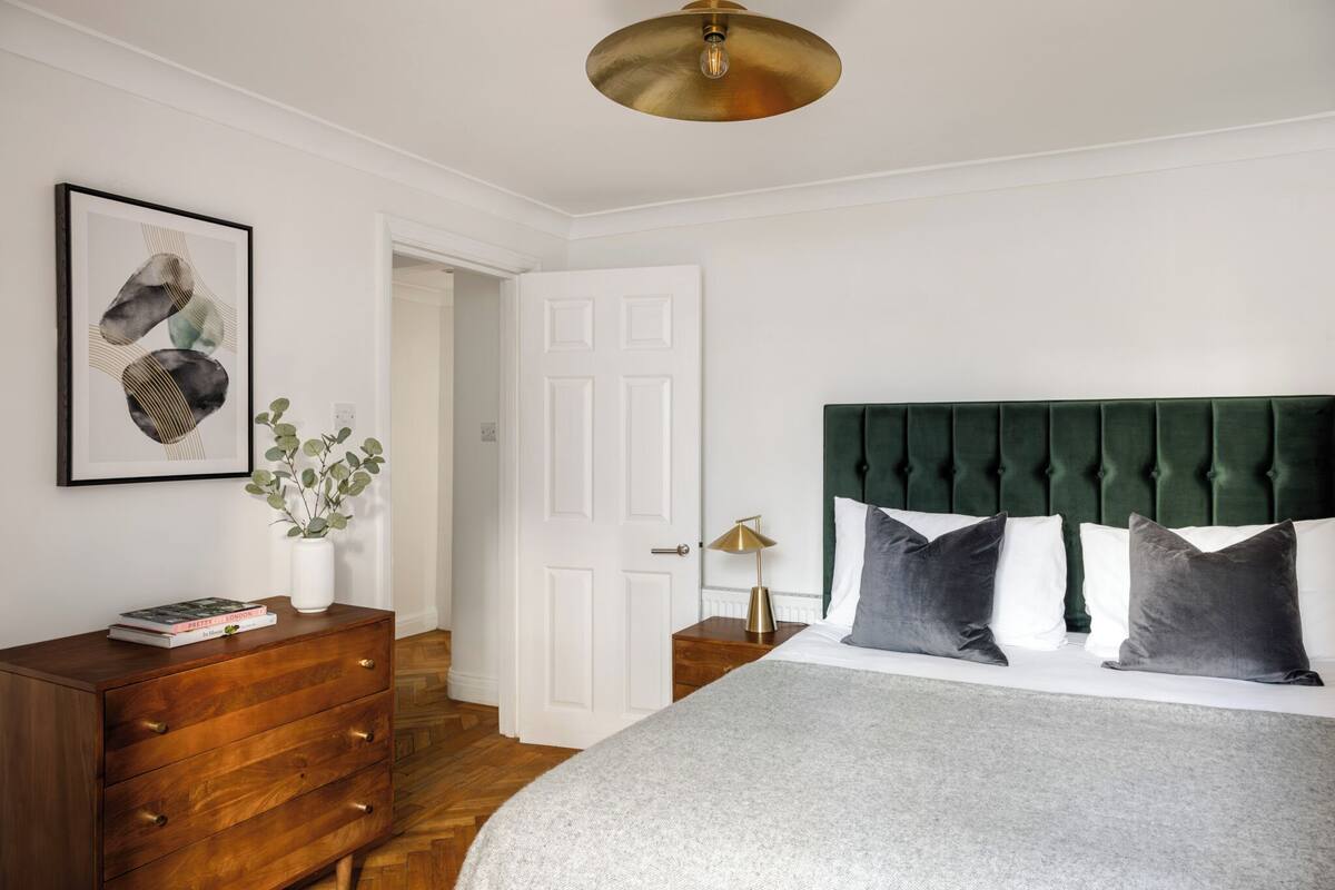 A bedroom with a green upholstered headboard, a wooden dresser, a framed abstract painting, and a bedside lamp.