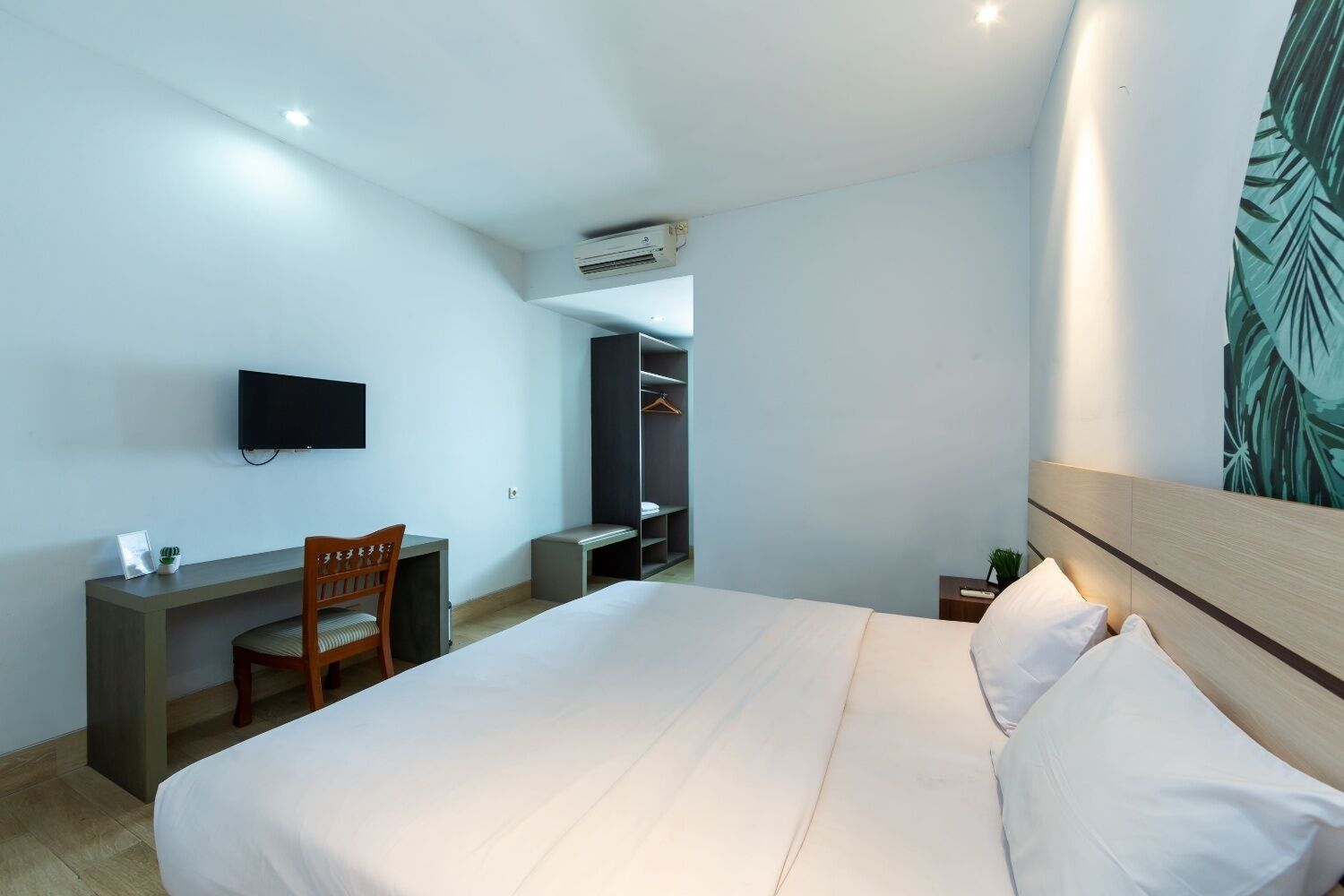 A hotel room with a bed, a desk with a chair, a TV, a wardrobe, and an air conditioning unit.
