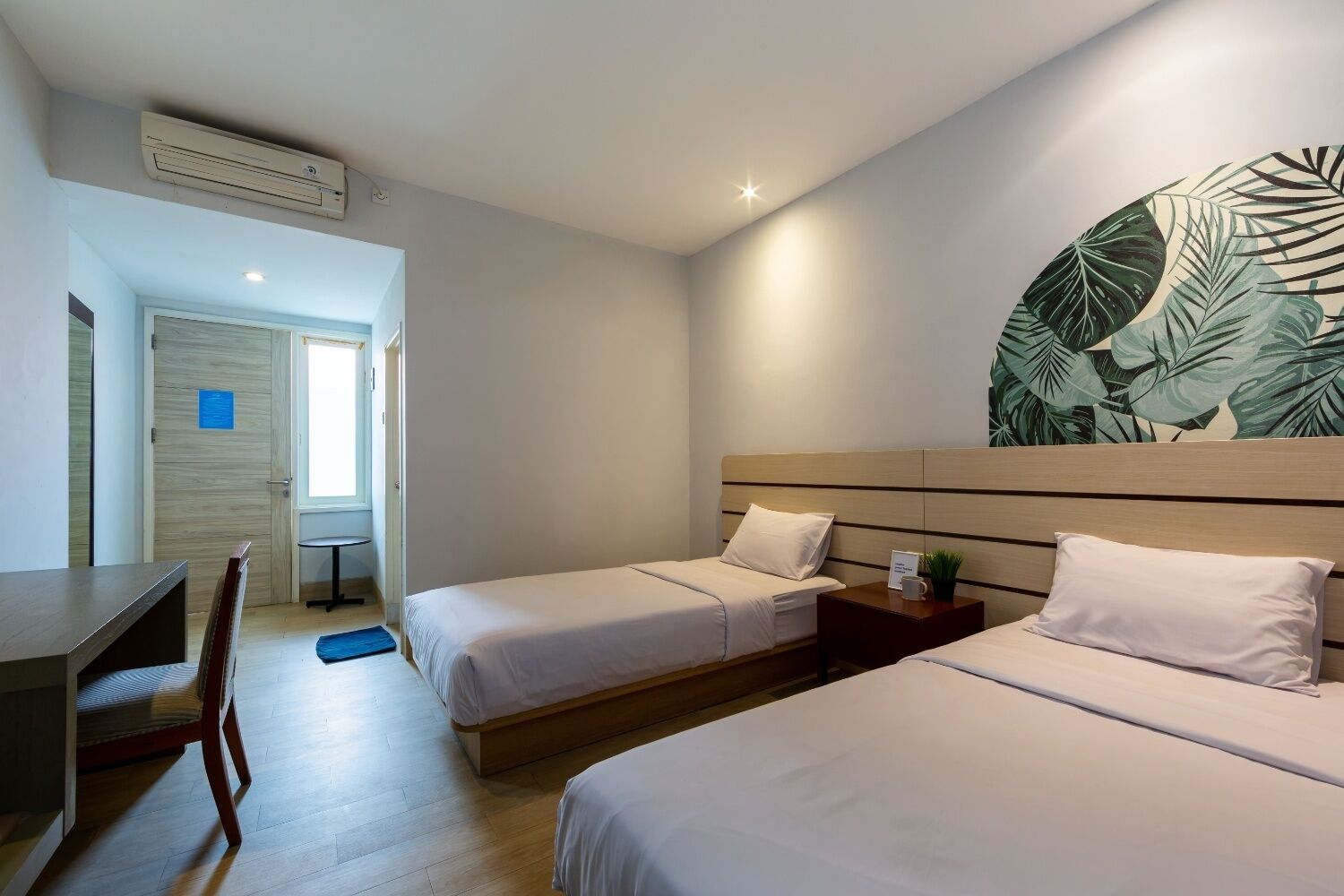 A hotel room with two beds, a desk, and a chair. There is a large window and a wall with a tropical leaf pattern.