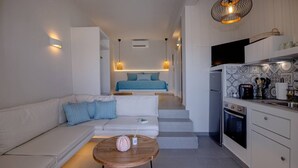 A compact living space with a sofa, a coffee table, a kitchen area with a refrigerator and stove, and a bed in the background. - Blue Pearl Suite in Tinos  (Tinos Town)