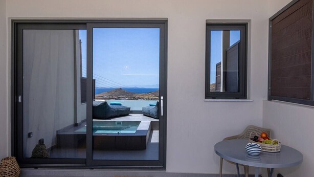 A balcony with a glass door, a small table with fruit, and a view of a pool and mountains. - Blue Pearl Suite in Tinos  (Tinos Town)