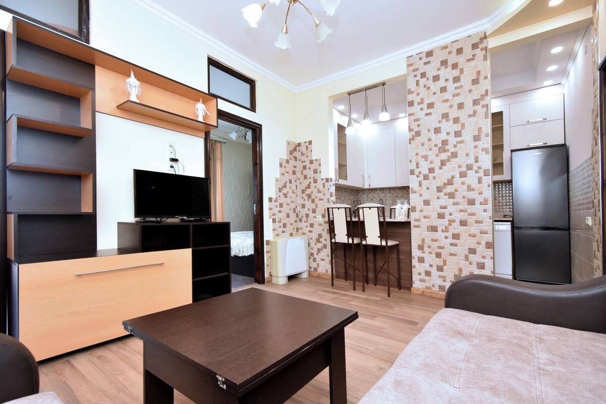 A modern living room with a flat-screen TV, wooden furniture, and a dining area with a kitchen in the background.