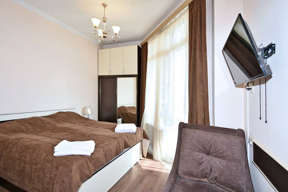 A bedroom with a bed, a TV mounted on the wall, a chair, and a window with curtains.