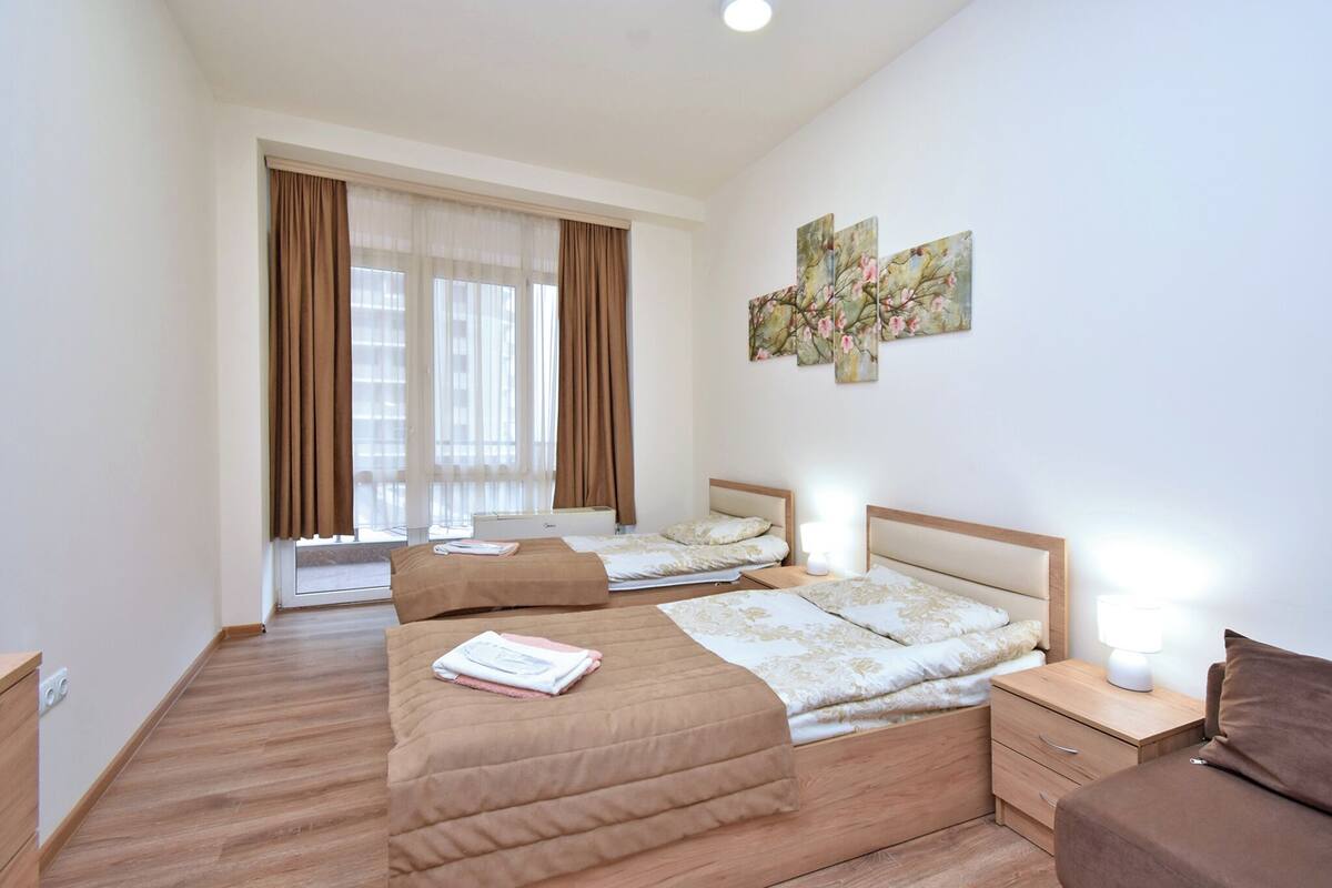 A hotel room with two beds, a large window with curtains, a bedside table with a lamp, and a wall with floral artwork.