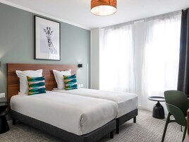 Habitación de hotel con dos camas, un cuadro enmarcado en la pared, una mesita de noche, una silla y un ventanal con cortinas translúcidas.
