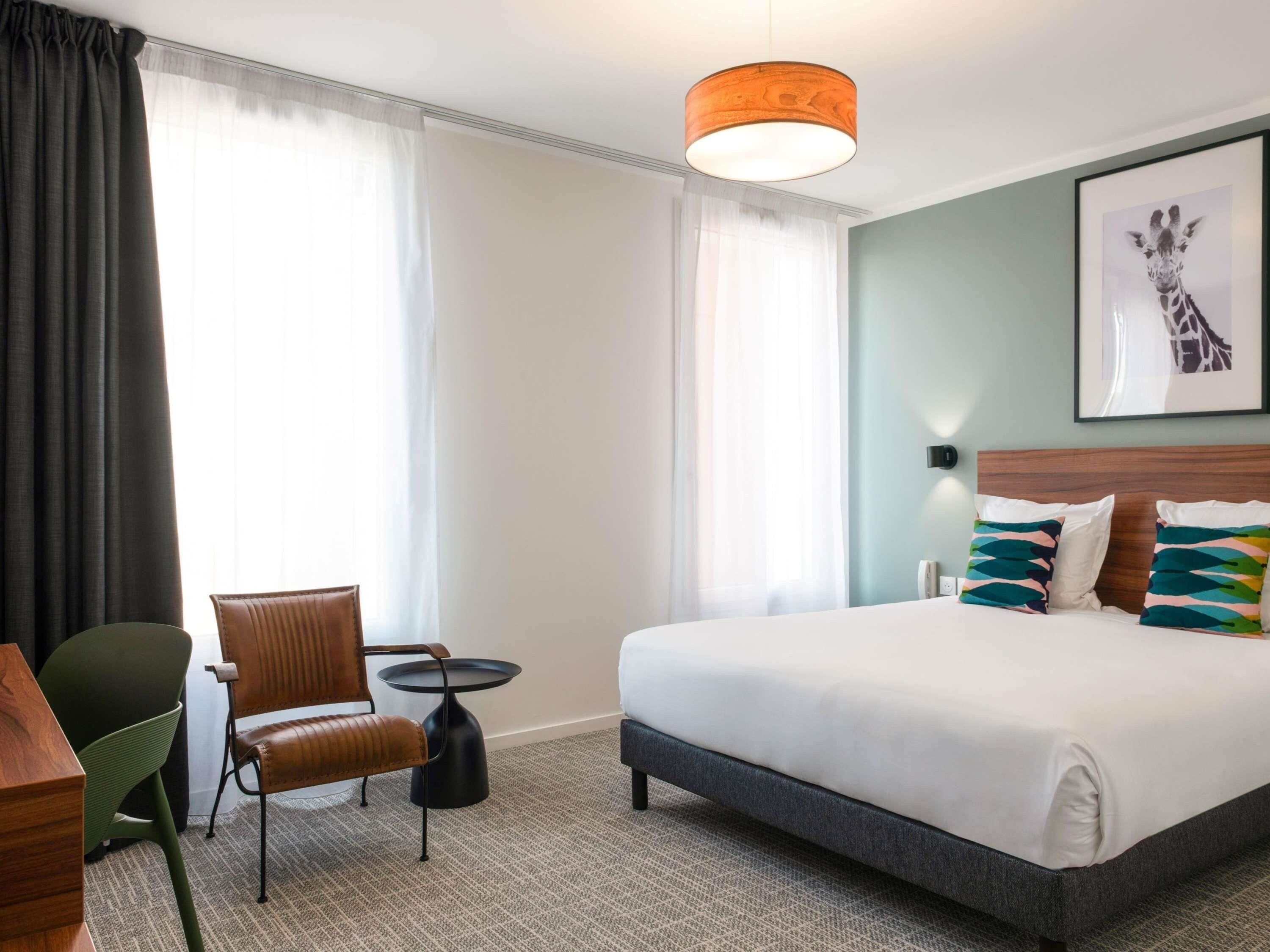 Een hotelkamer met een bed, een stoel, een klein tafeltje en een groot raam met gordijnen.