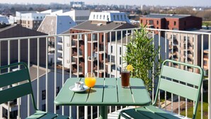 Un balcón con una mesa, sillas y una bebida. El balcón tiene vistas a un paisaje urbano con varios edificios.