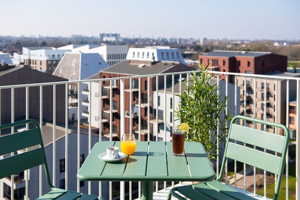Un balcón con una mesa, sillas y una bebida. El balcón tiene vistas a un paisaje urbano con varios edificios.
