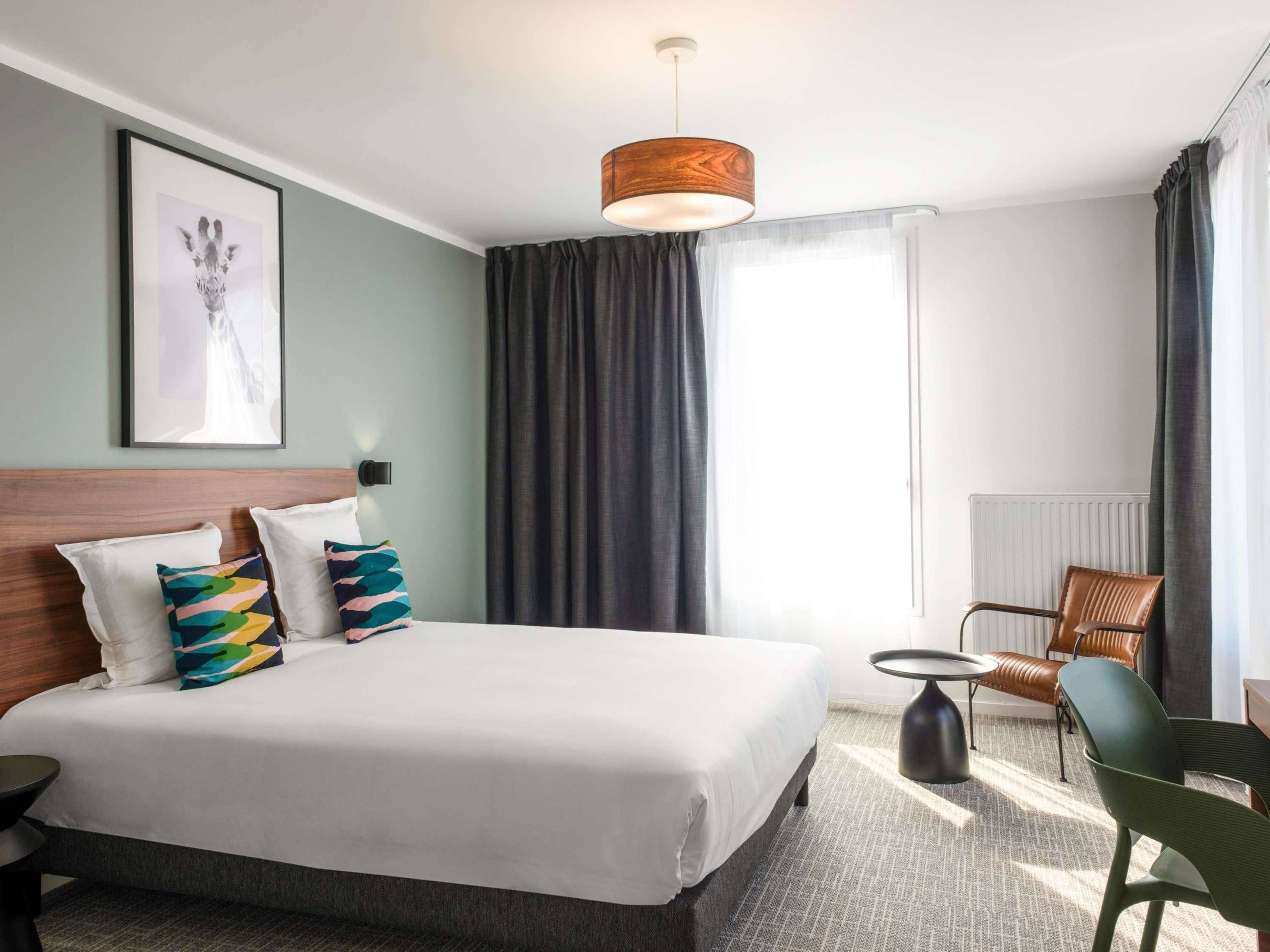 A hotel room with a large bed, a side table, a chair, a framed picture on the wall, and a window with curtains.