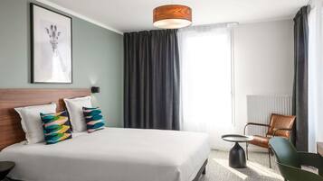 A hotel room with a large bed, a side table, a chair, a framed picture on the wall, and a window with curtains.