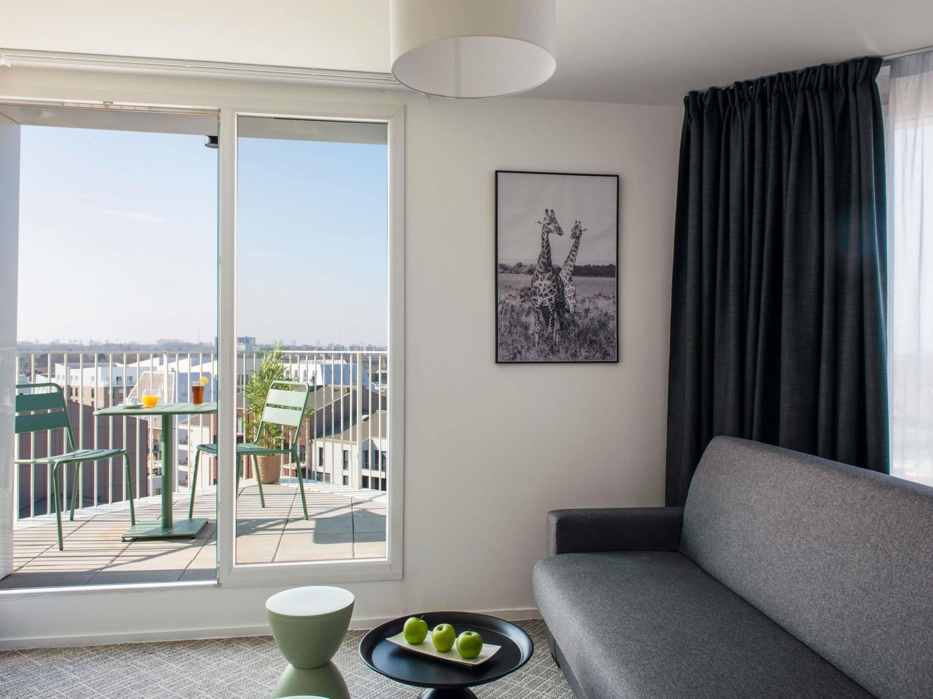 A modern living room with a grey sofa, a dining table with chairs, a balcony with a view of buildings, and a framed picture on the wall.