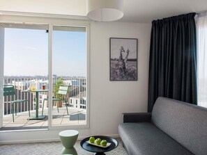 A modern living room with a grey sofa, a dining table with chairs, a balcony with a view of buildings, and a framed picture on the wall. - Aparthotel Adagio Access Lille Villeneuve-d'Ascq (Villeneuve-d'Ascq)