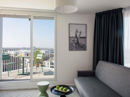 A modern living room with a grey sofa, a dining table with chairs, a balcony with a view of buildings, and a framed picture on the wall.