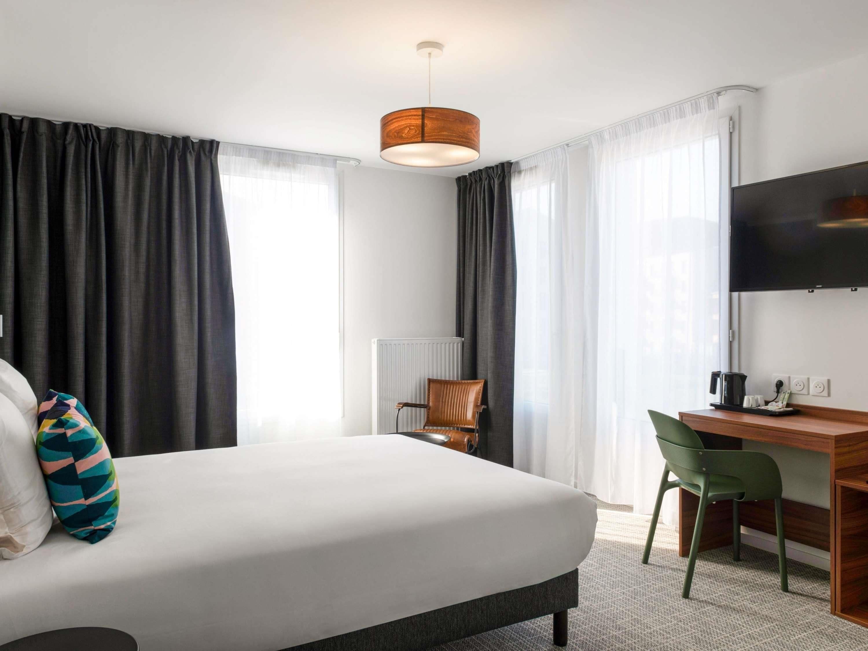 Habitación de hotel con cama, escritorio, silla, televisión y ventana con cortinas.