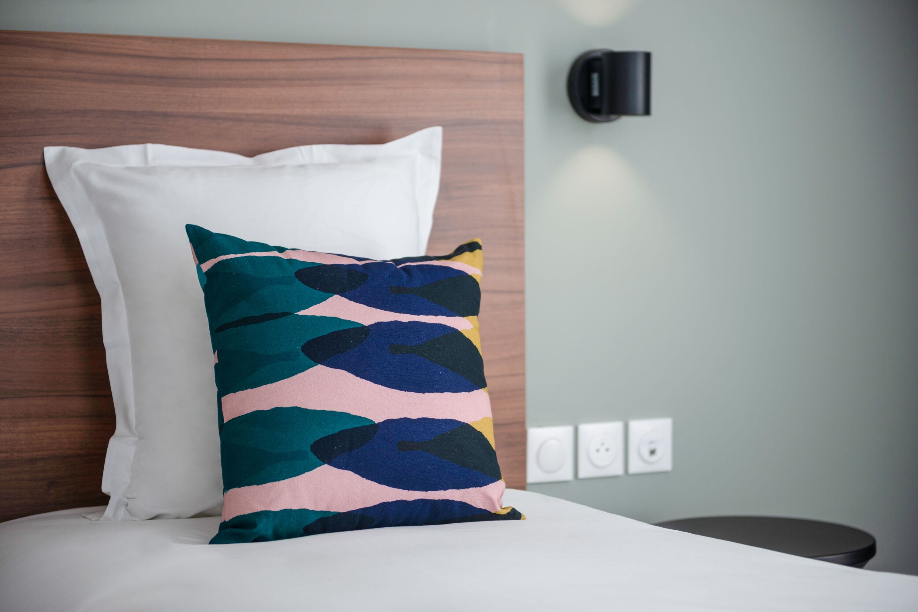 A bed with a white pillow and a colorful pillow, a wooden headboard, a black wall-mounted lamp, and a round table.