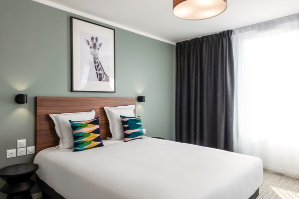A neatly made bed with white bedding and two colorful pillows, a framed giraffe picture on the wall, and a dark curtain covering a window. - Aparthotel Adagio Access Lille Villeneuve-d'Ascq (Villeneuve-d'Ascq)