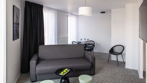 Una sala de estar moderna con un sofá gris, una mesita con una bandeja de manzanas, una zona de comedor con una mesa y sillas, y una silla negra.