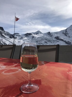 Ein Glas Weißwein auf einem Tisch mit roter Tischdecke, vor einer Kulisse aus schneebedeckten Bergen und einer Flagge.